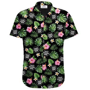 A short-sleeve button-up camp shirt with a black background and an all-over tropical print of green monstera and palm leaves, pink hibiscus flowers, and white plumeria. The shirt features customizable circular logo placeholders with the text 'YOUR LOGO HERE' and 'YOUR PHOTO HERE'.
