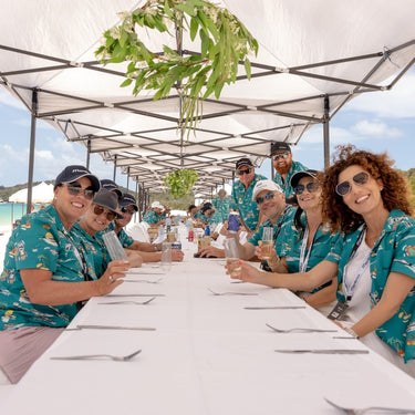 Group of people in matching teal shirts sitting at a long table under a white tent with greenery.